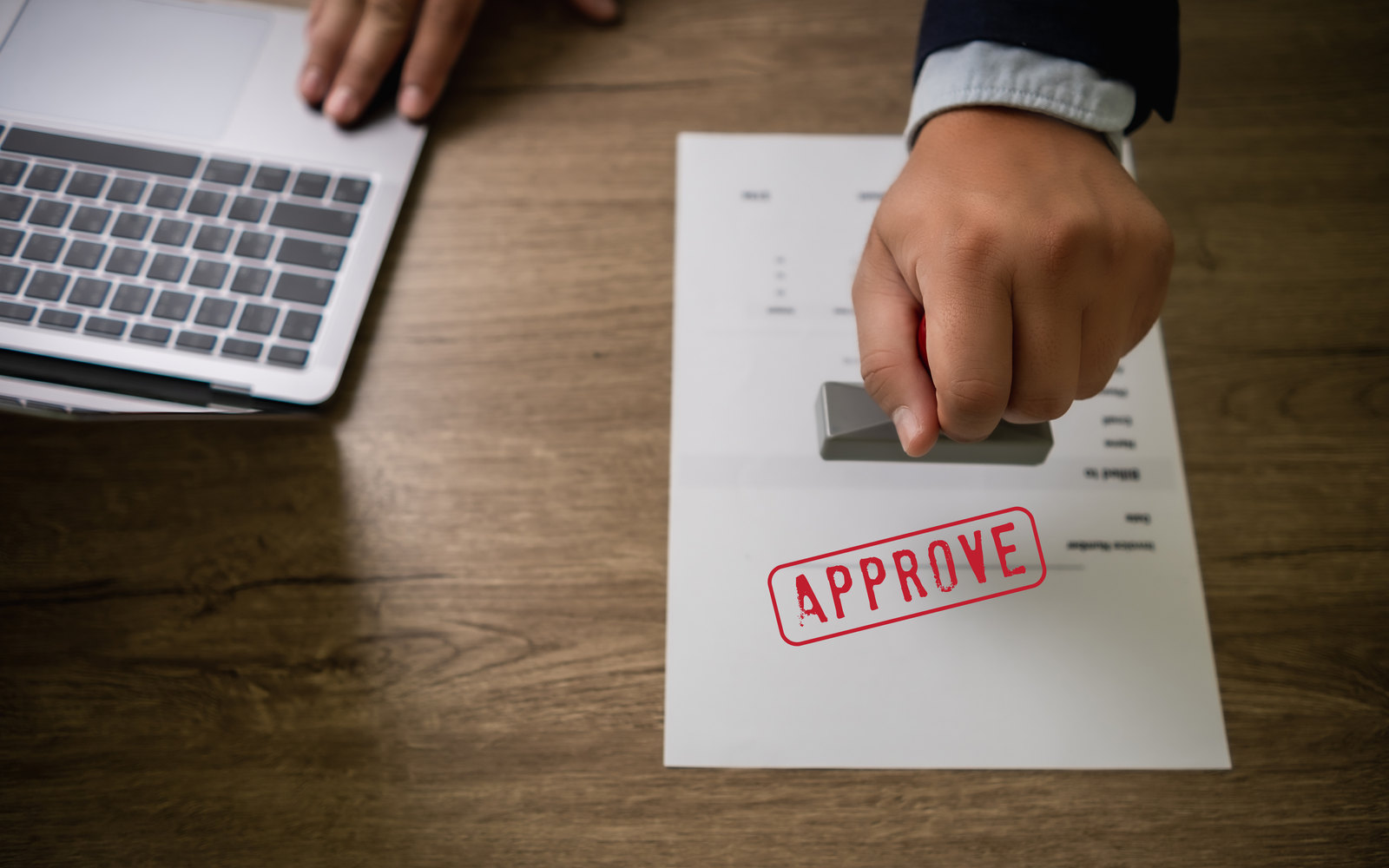 A hand pressing a red APPROVE stamp onto a document beside a laptop