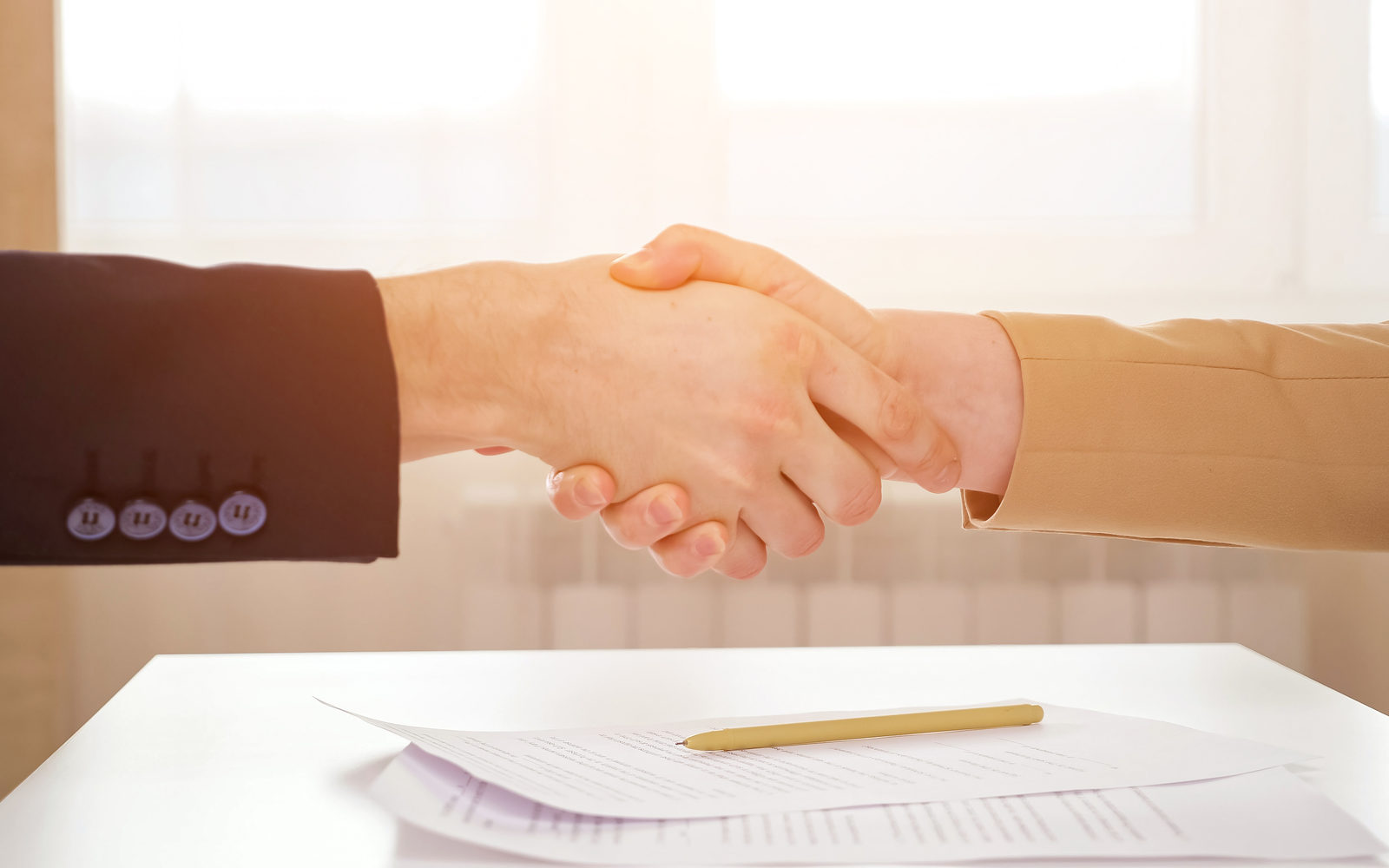 Two hands shaking over a contract and pen on a white table in warm window light