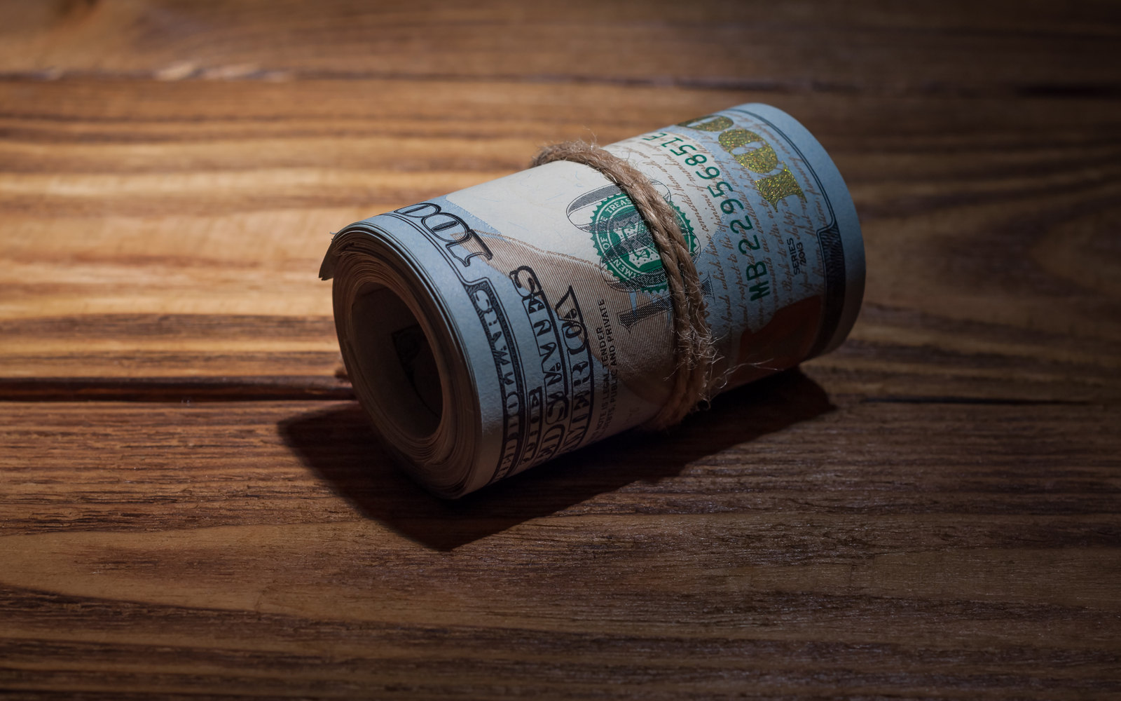 A tight roll of hundred-dollar bills tied with twine on a dark wooden table
