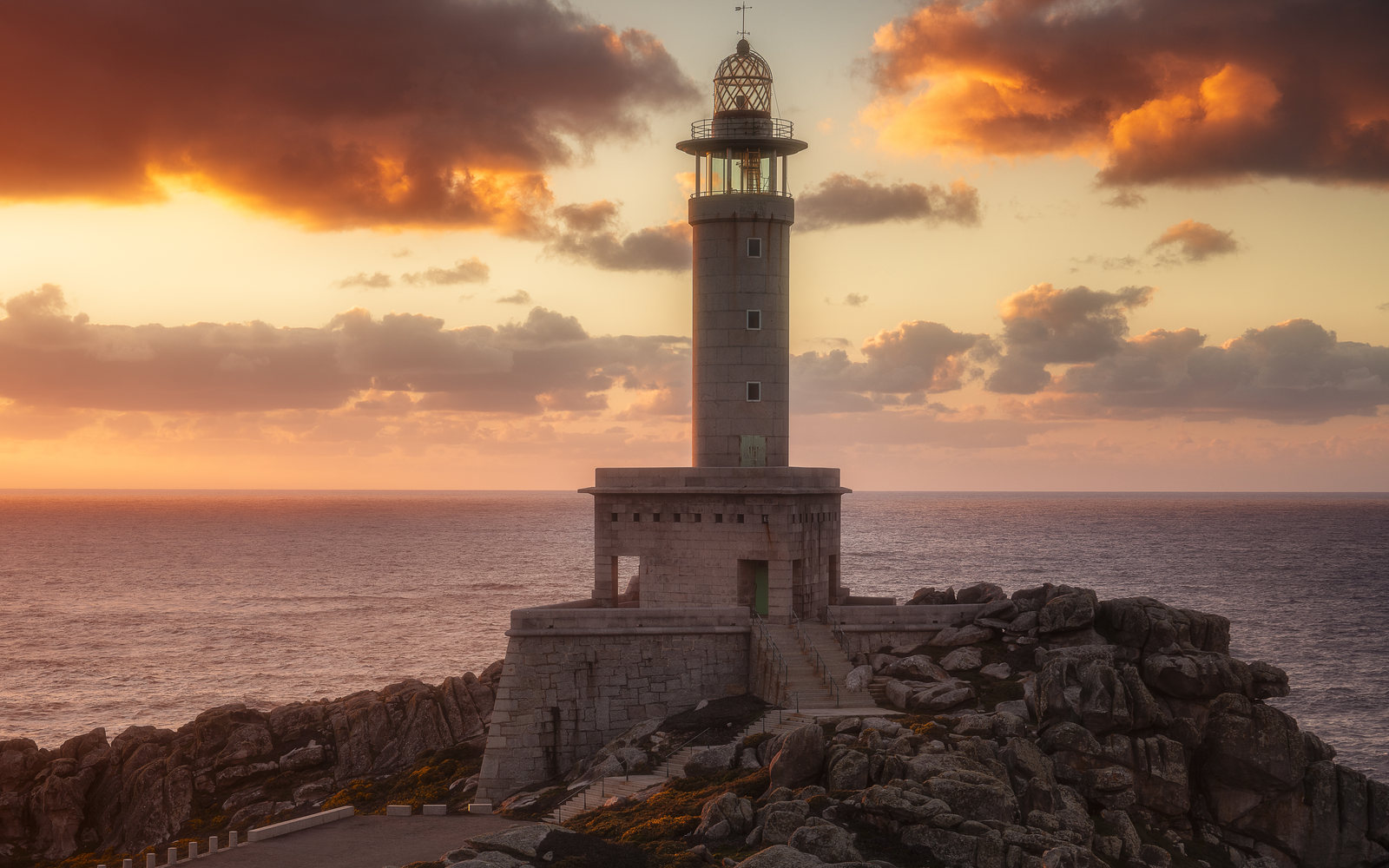 Stone lighthouse on a rocky headland under a warm, dramatic sunset sky