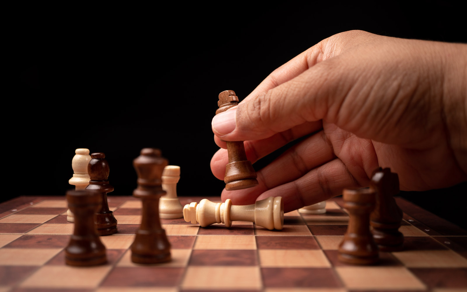 A hand making a decisive chess move on a wooden board against a black background