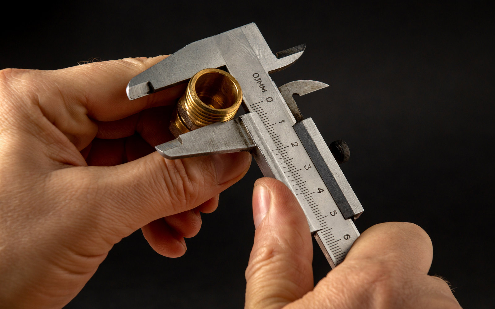 Hands measuring a brass fitting with a steel caliper against a black background