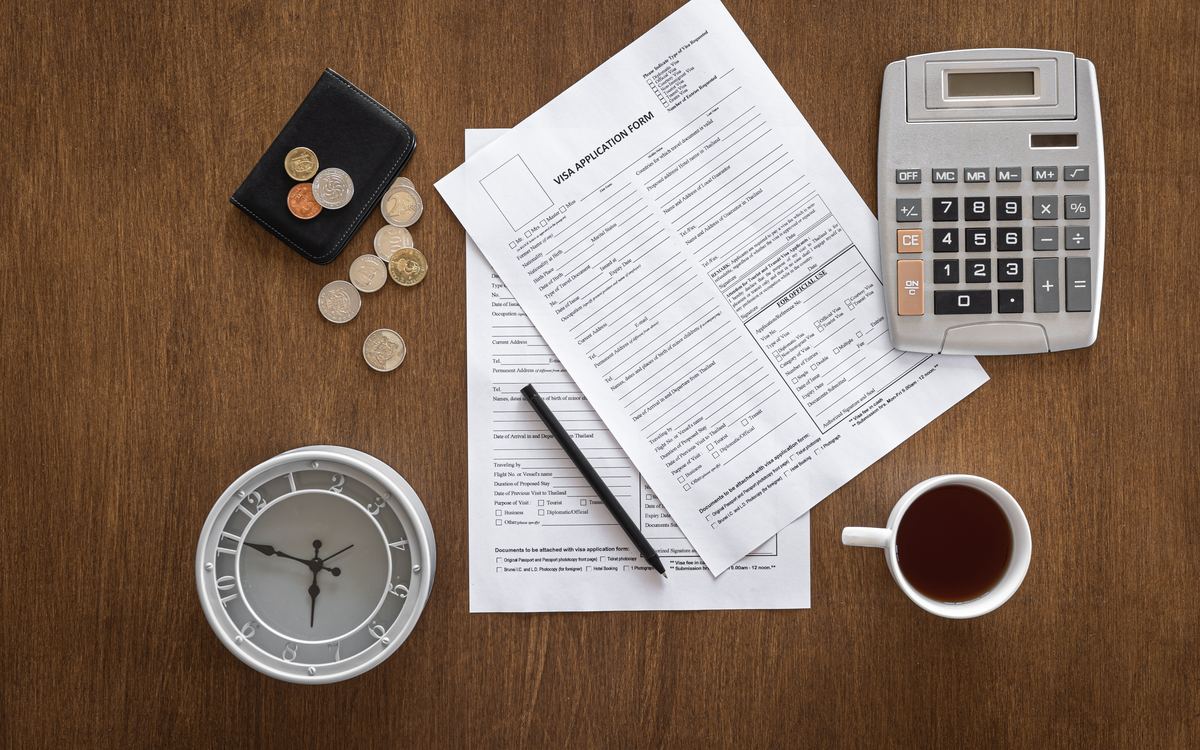 Application forms, calculator, clock, coins and tea cup arranged on a wooden desk